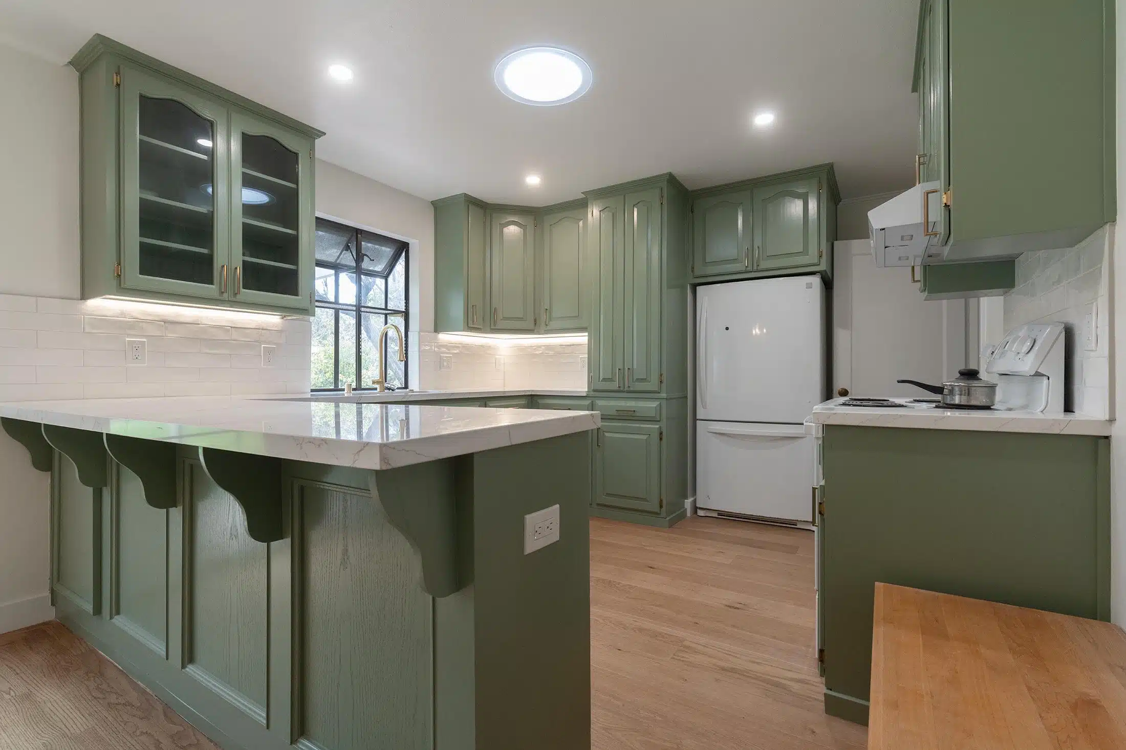 Sage green kitchen with a corbelled peninsula, glass-front uppers and a skylight